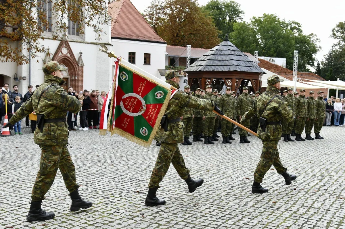 Żołnierze w kamuflażu maszerują z sztandarem podczas uroczystości wojskowej na brukowanym rynku, w tle stoją w szeregu koledzy i zgromadzeni widzowie.