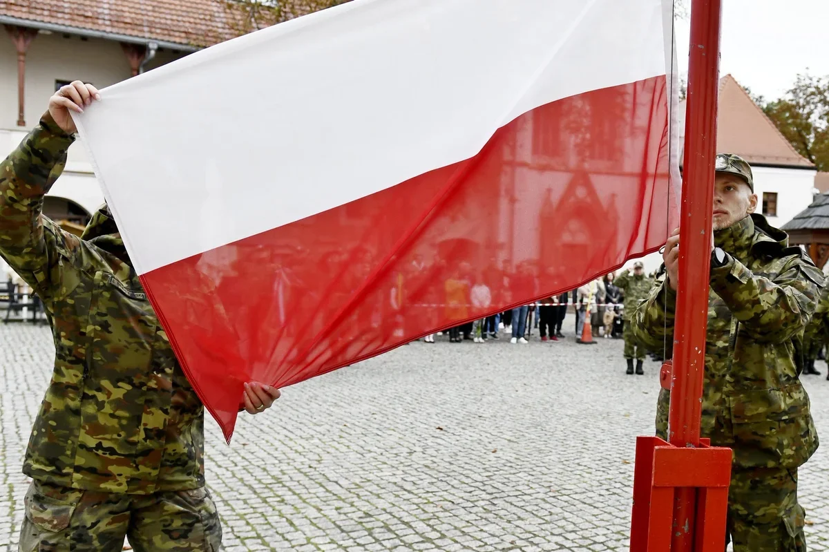 Żołnierze w kamuflażu uczestniczą w ceremonii wciągania polskiej flagi na maszt na brukowanym rynku miejskim.