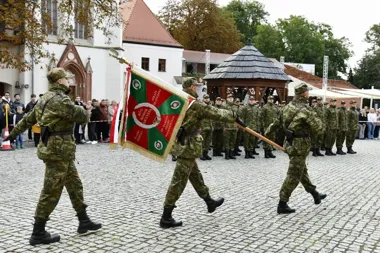 Żołnierze w kamuflażu maszerują z sztandarem podczas uroczystości wojskowej na brukowanym rynku, w tle stoją w szeregu koledzy i zgromadzeni widzowie.