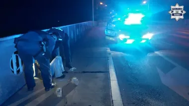 Policjanci interweniują w nocy na moście przy chodniku obok radiowozu z włączonymi niebieskimi światłami.