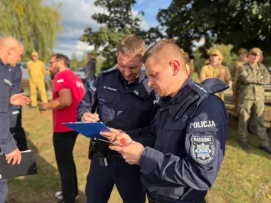 Policjanci z komendy powiatowej w Raciborzu wypełniają dokumentację podczas działań na terenie trawiastym, w tle stoją osoby w mundurach i cywilni uczestnicy.